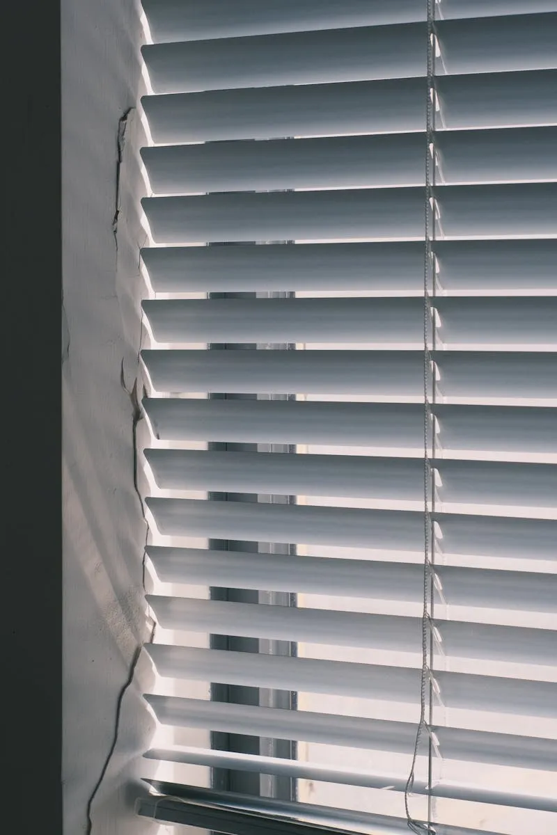 Halb geöffnete weiße Lamellen-Jalousie vor einem Fenster, von innen fotografiert
